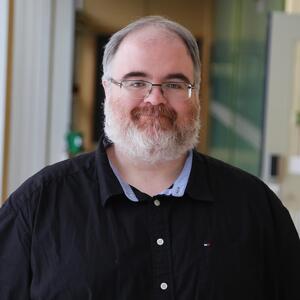 Steven grins at the camera. He is wearing a black collared button up shirt and glasses. He has short salt & pepper hair and a white beard.