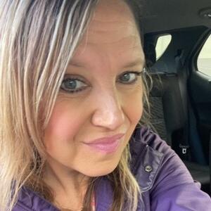 April looks at the camera. She is wearing a purple jacket. She has straight blond hair.