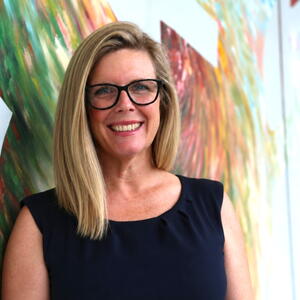 A woman with glasses smiles in front of art work 