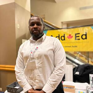 Akomaye Undie poses in front of a poster for the Add*Ed research group wearing a white button down.
