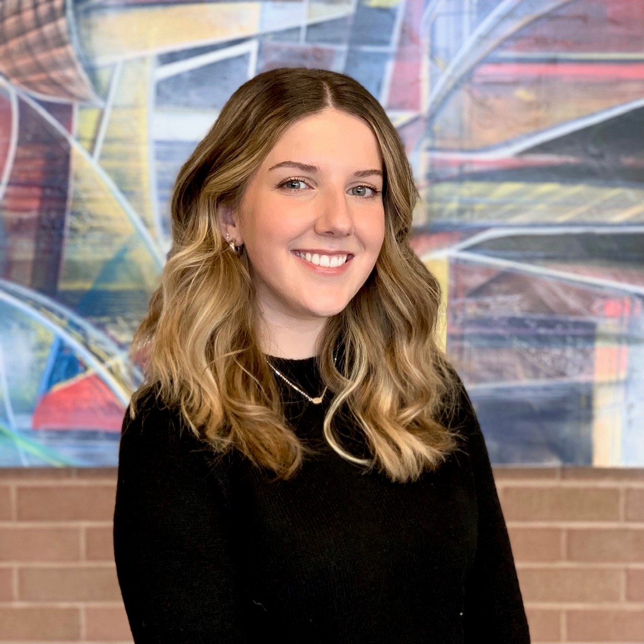 Andrea, who has medium length blonde hair, smiles at the camera wearing a black shirt. There is a multicoloured painting on a brick wall in the background.