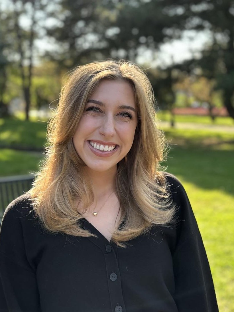 Erica smiles broadly at the camera. With long blond hair, there is a park in the background.