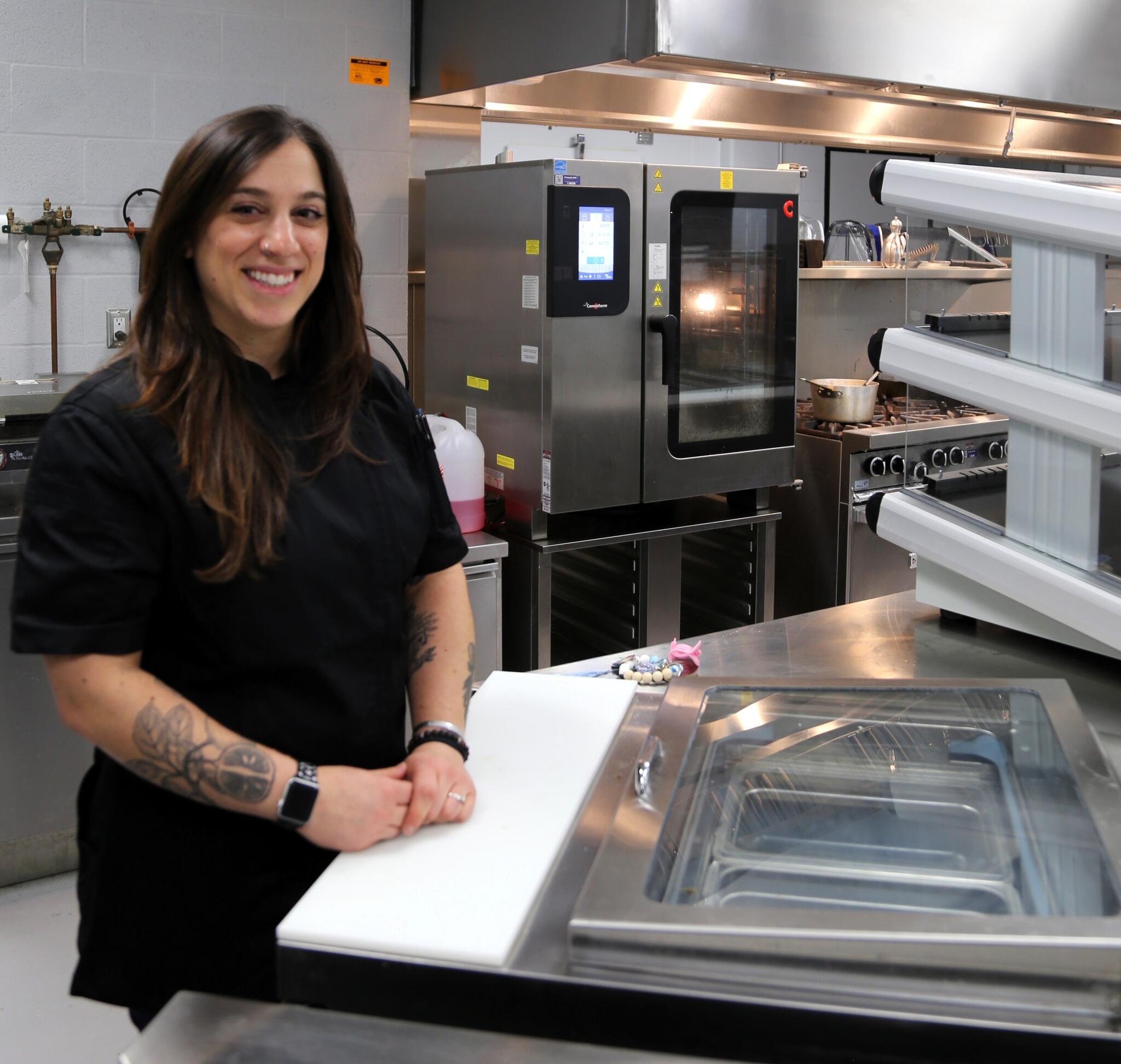 Grace smiles at the camera with dark hair and chefs jacket on. She is in a kitchen space with lots of stainless steal surfaces