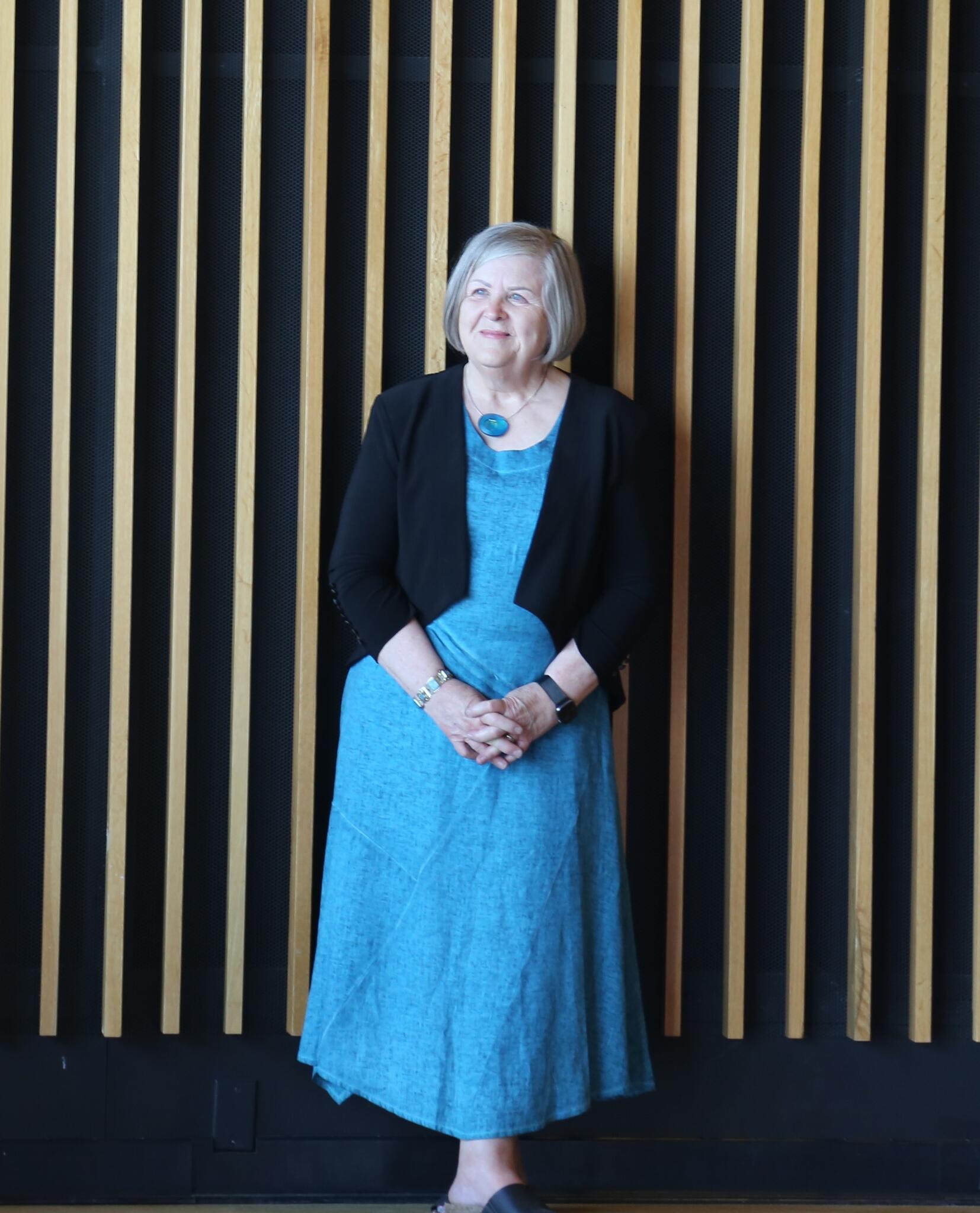 Rebecca leans against a wall wearing a blue dress and a black cardigan. She is smiling and looking to the right of the camera