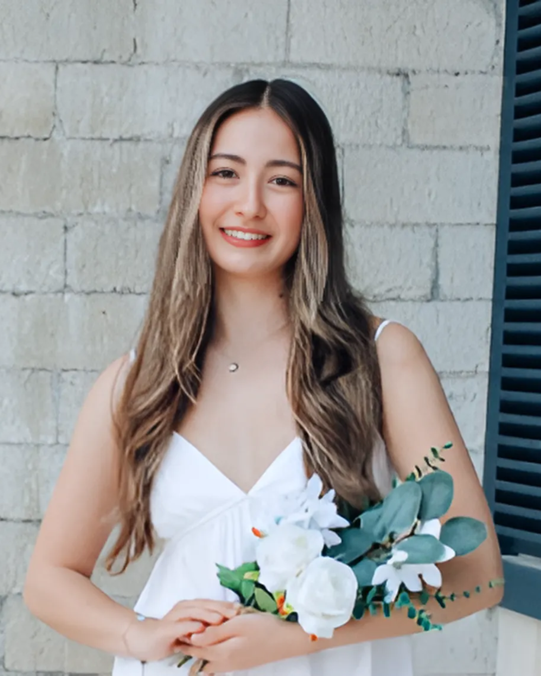 Mya smiles at the camera wearing a white dress and holding flowers. 