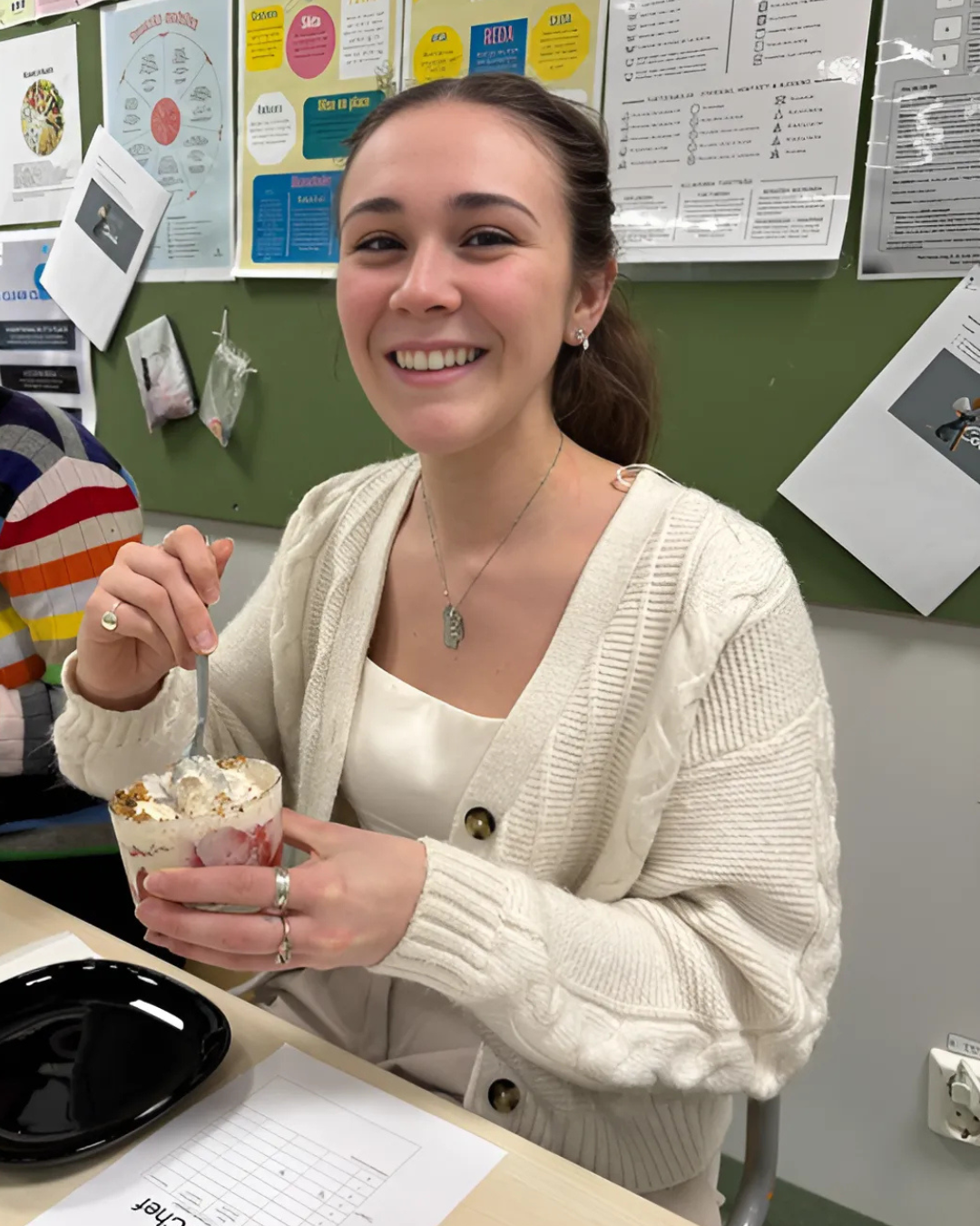 Sarah is in a classroom with a quart of ice cream and a spoon in her hand. She is smiling broadly at the camera.