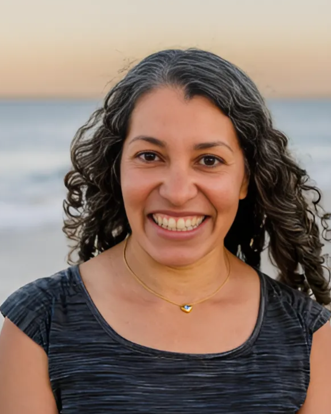 Nerissa, standing in-front of a beach smiles broadly. Narissa has shoulder length curly dark hair and is wearing a grey t-shirt