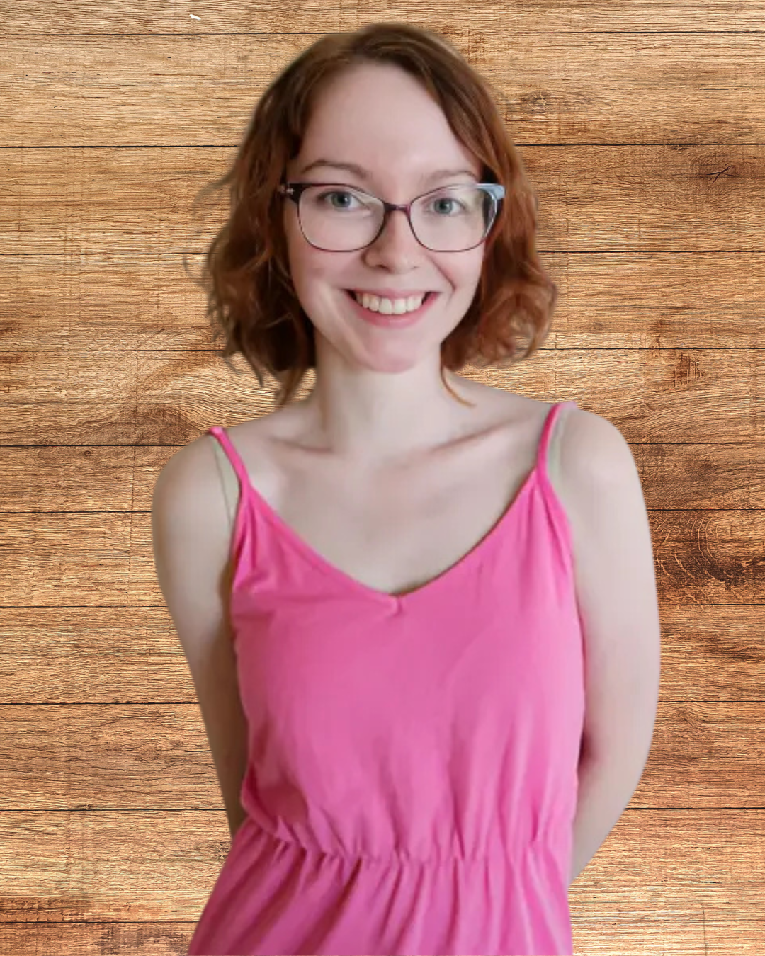 Claire, wearing a pink dress is smiling at the camera. She has red, chin length hair and is wearing glasses