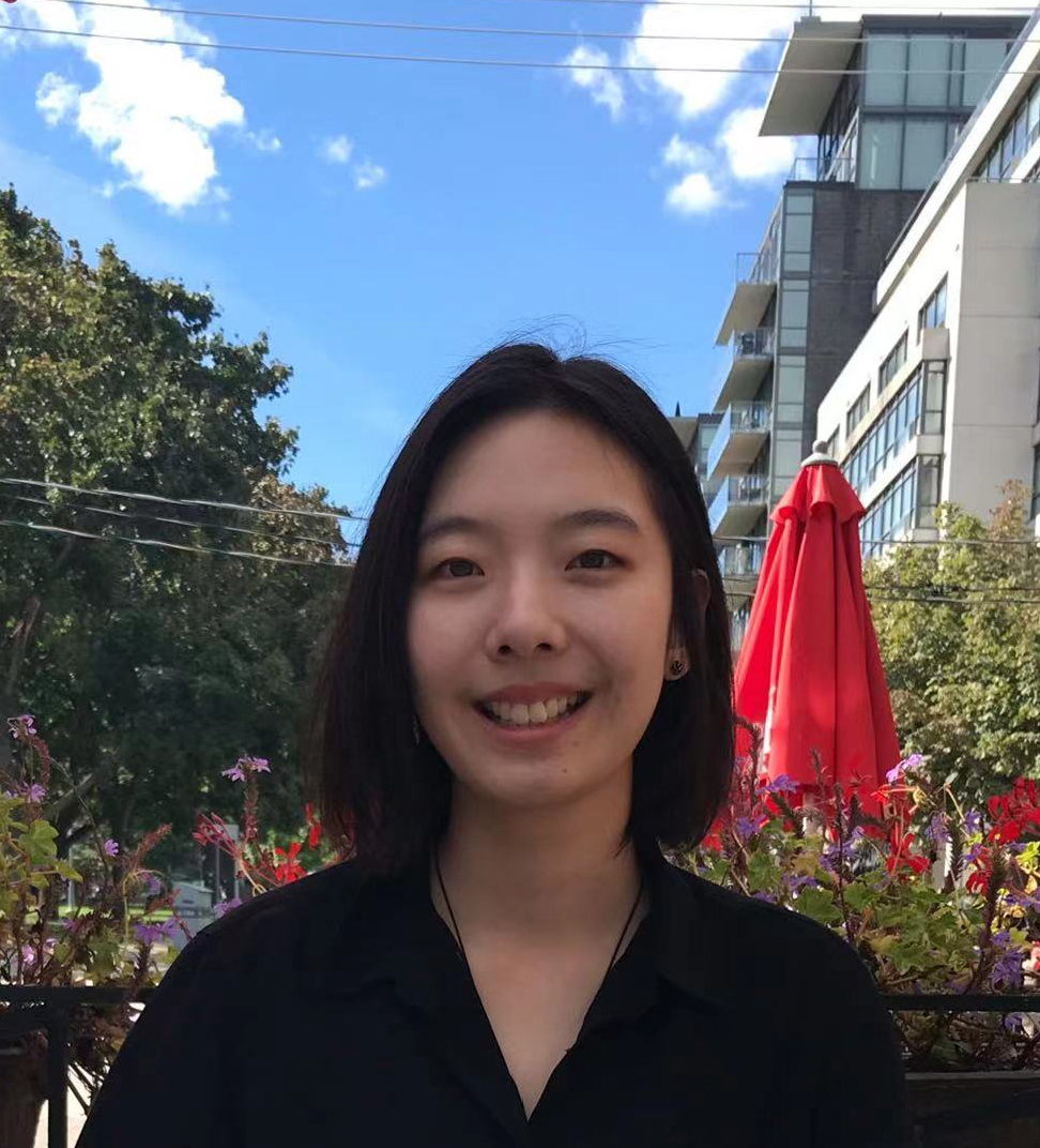 Jianing, smiles at the camera with blue skies and an outdoor cafe with red umbrellas in the background.