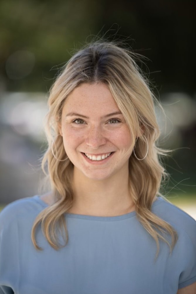 Julia, with long blond hair smiles at the camera wearing a light blue shirt, the background is blurry with green and golden light.