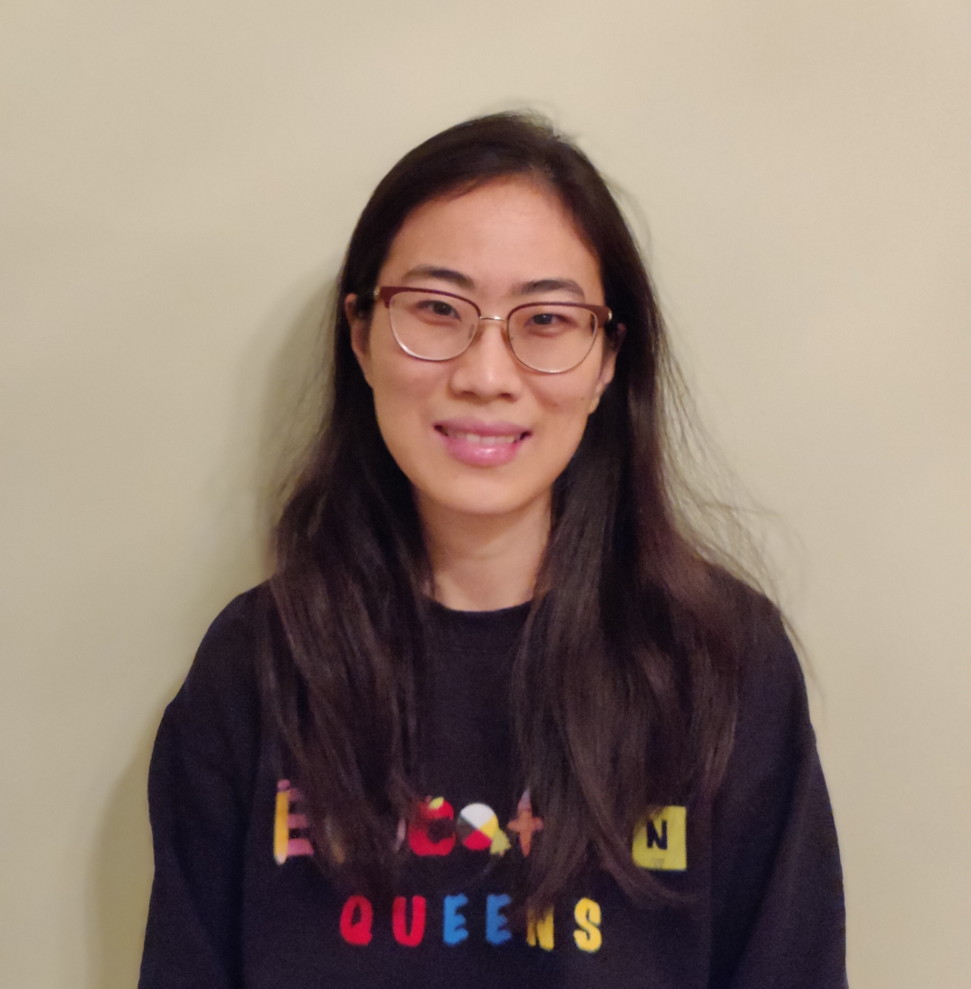 Laura smiles at the camera with long brown hair, standing against a beige background, wearing a Queen's Education shirt