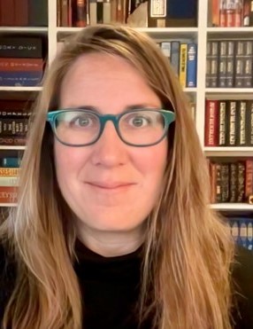 Lydia smiles at the camera with green glasses, long blond hair and a bookshelf in the background