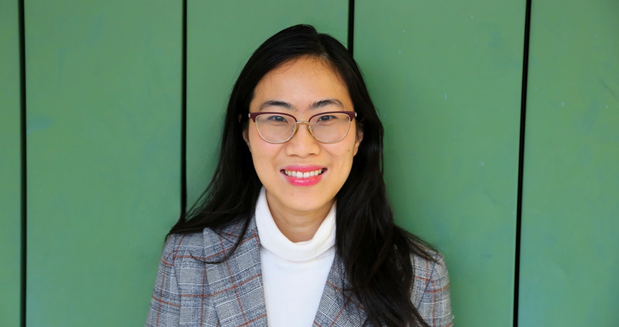 An asian woman with long black hair wearing classes, a white turtle neck and a plaid blazer