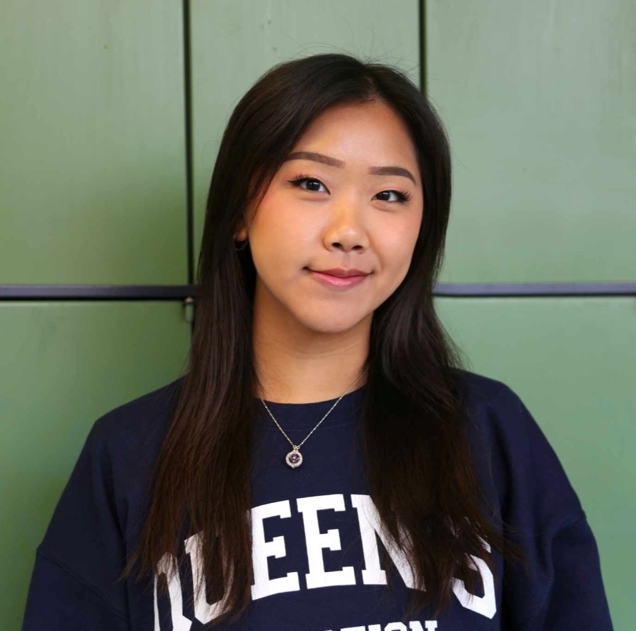 Rachel smiles against a green background wearing a Queen's Education shirt. Rachel's hair is long and black.