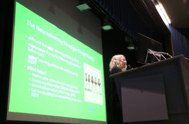 A speaker stands at a podium with a large screen with a bright green slide on it is in the background