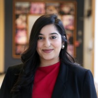 Sunaira has shoulder length black hair and smiles at the camera wearing a red shirt and black jacket 