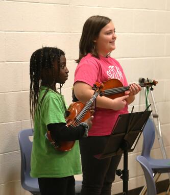Two students stand holding violins in a gym.