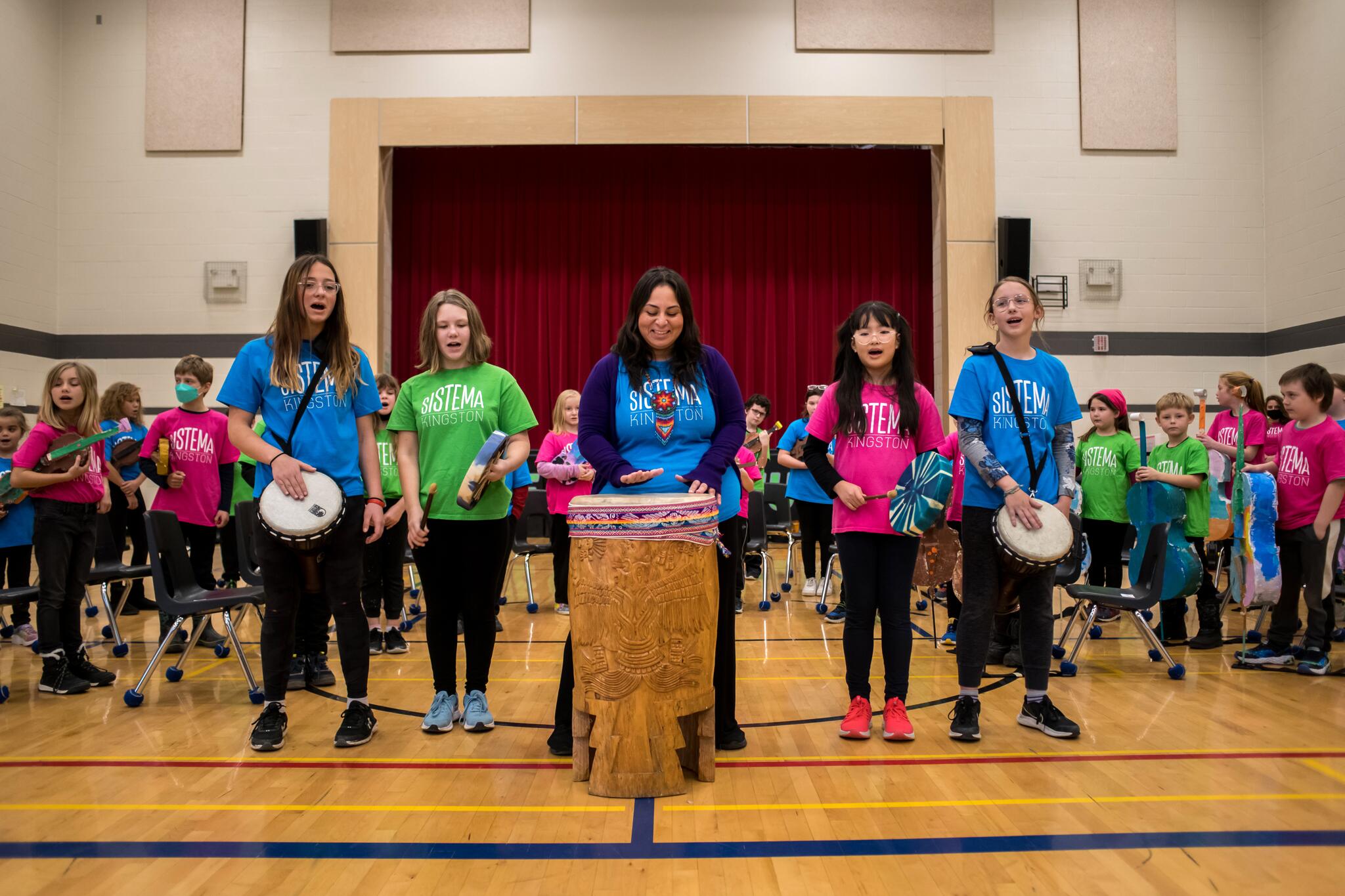 Students in colourful t-shirts stand in a semi-circle with instruments. They are led by a teacher on a large wooden drum that stands in the middle.