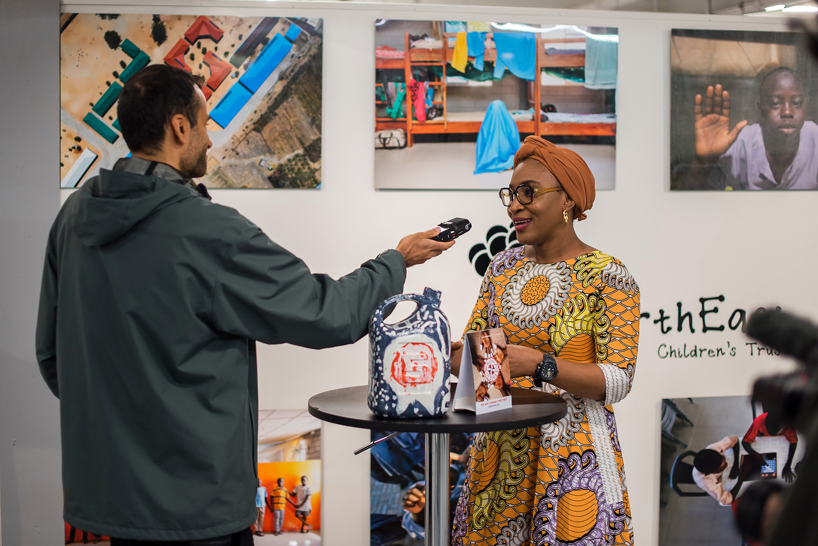 A woman in a colourful dress and head piece smiles while a man outstretches his arm to interview her.