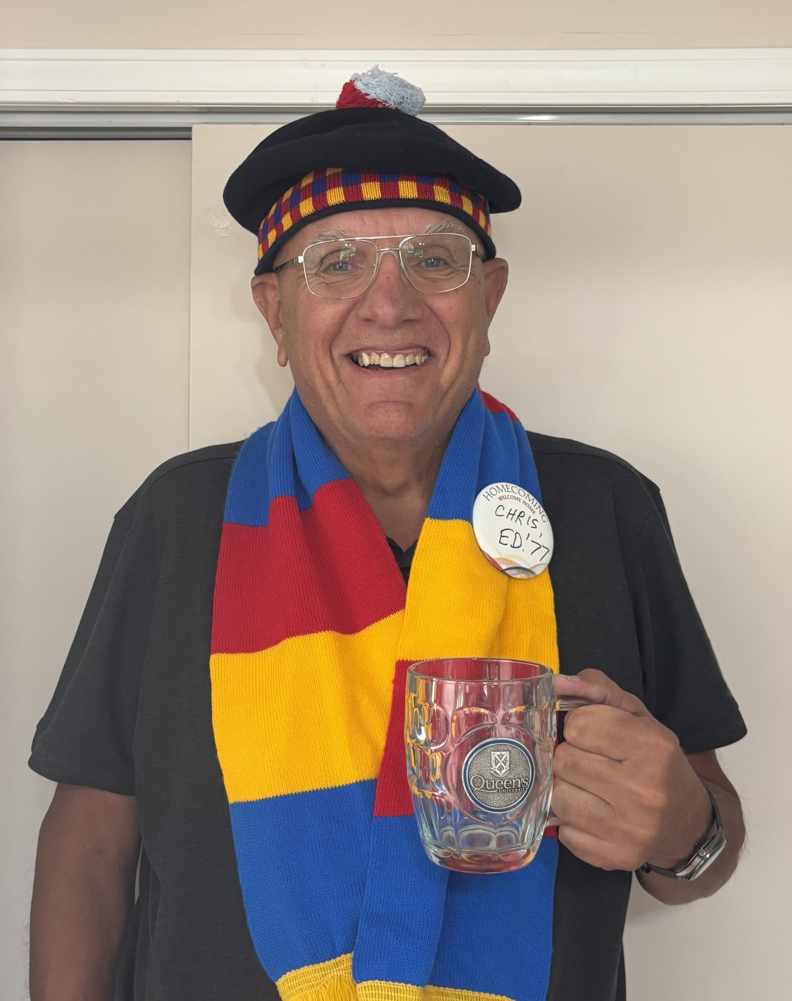 Chris Bobra smiles holding a Queen's mug and wearing a Queen's tam and scarf in a photo from 2025.