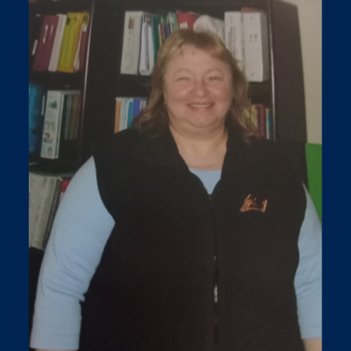 Connie Rosarios smiles wearing a vest and blue shirt standing in front of a bookshelf.