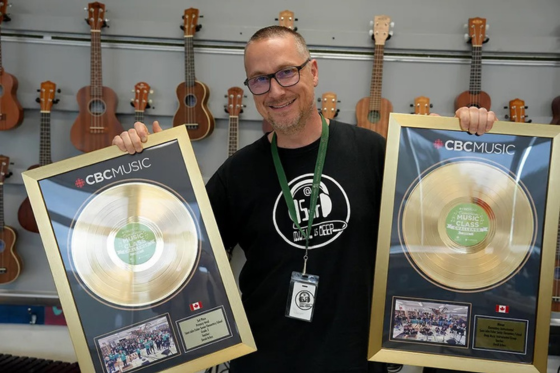 Dave Arless holds two gold record CBC plaques in front of a wall of ukuleles. He wears glasses and a black tshirt.