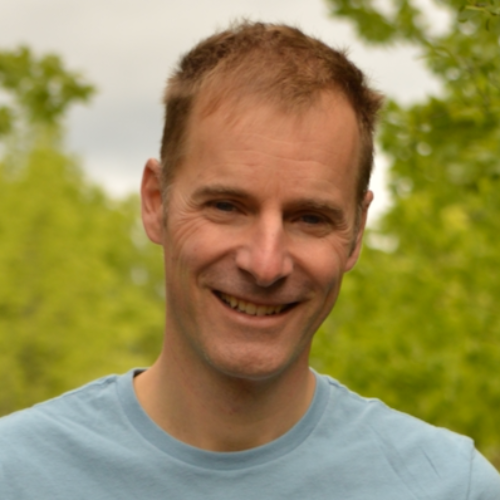 David Rankine smiles wearing a light blue shirt outside in front of green trees.