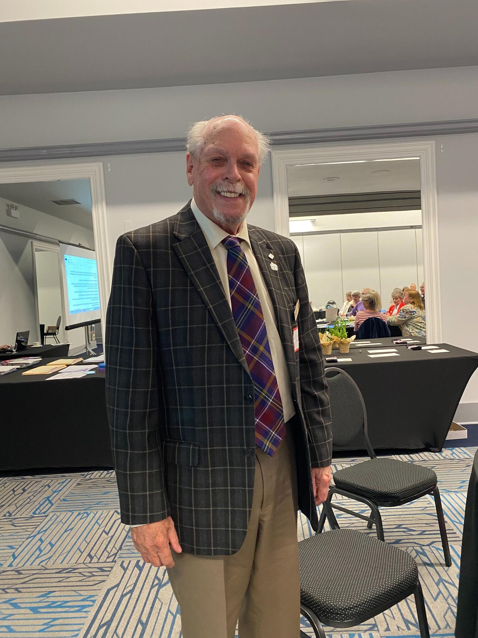 Dennis Quinn wears a suit and stands in a hall with chairs.