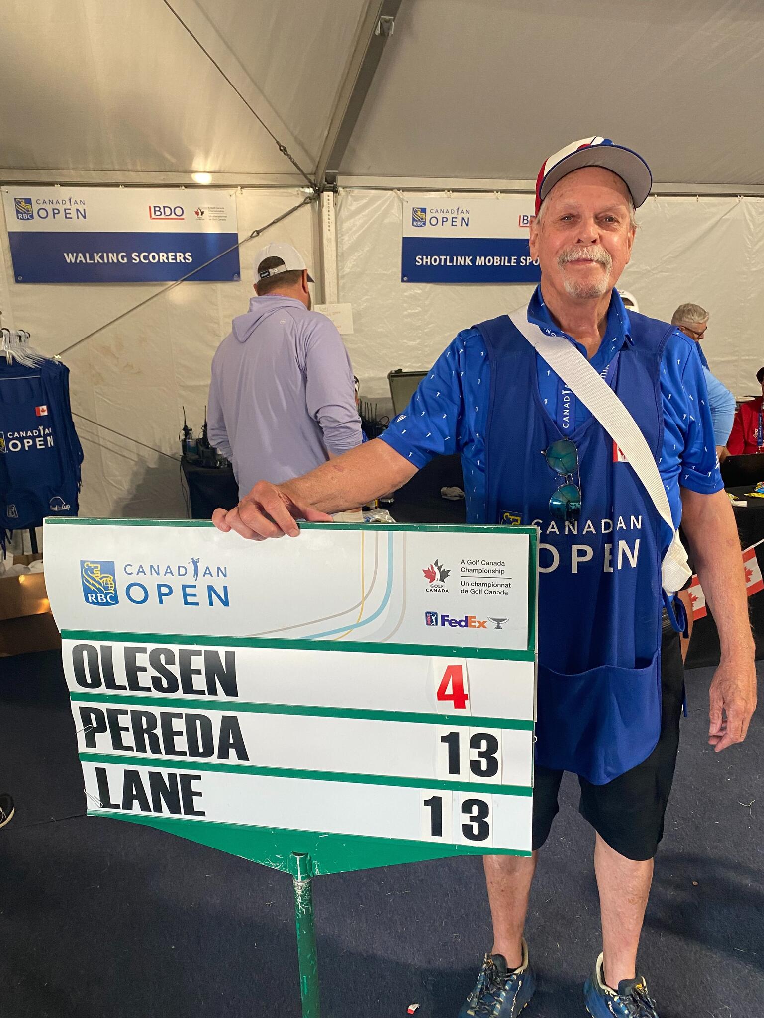 Dennis Quinn smiles wearing staff apparel for the Canadian Open and holds a placard with the last names of several athletes.