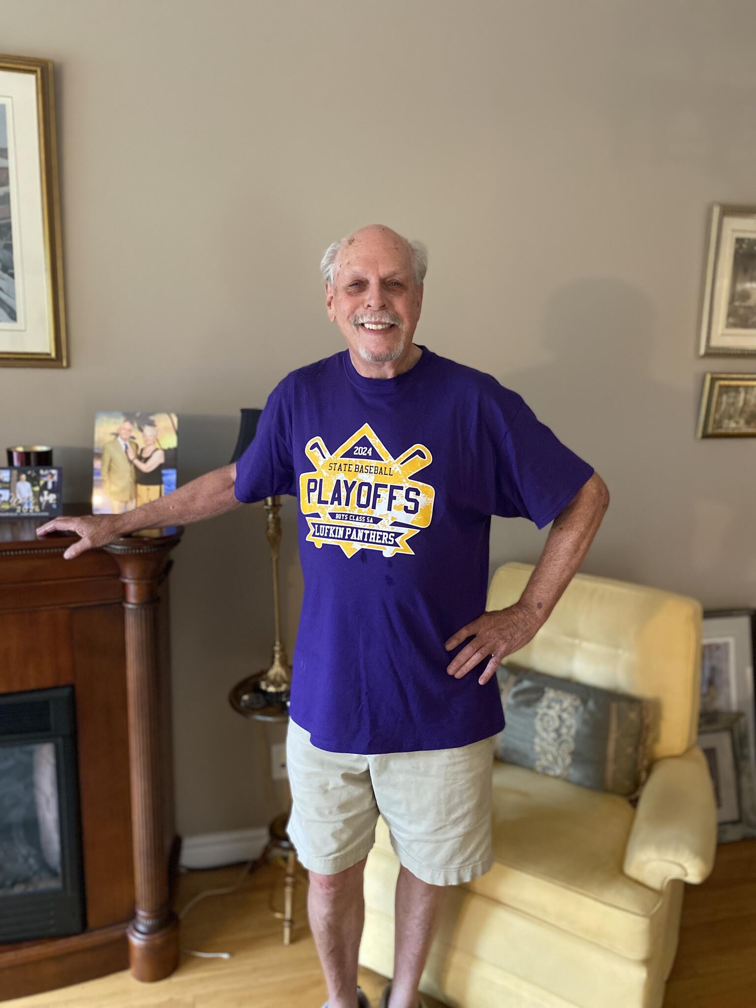 Dennis Quinn smiles wearing a baseball playoffs shirt and standing in a living room in front of a chair in 2025.