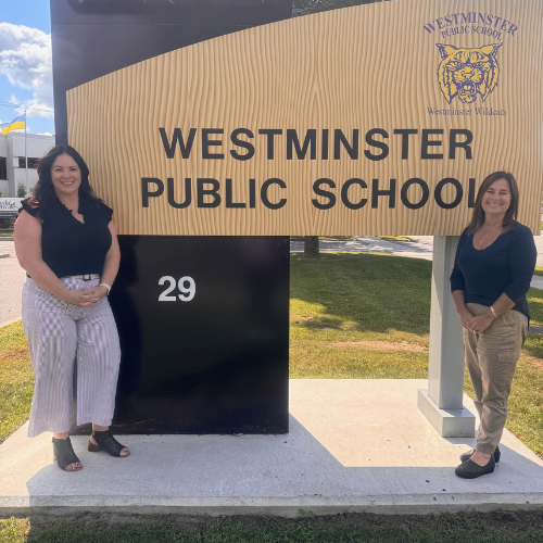 Jennifer McMaster smiles standing to the right side of a sign for Westminster Public School. She poses with another woman standing to the left of the sign.