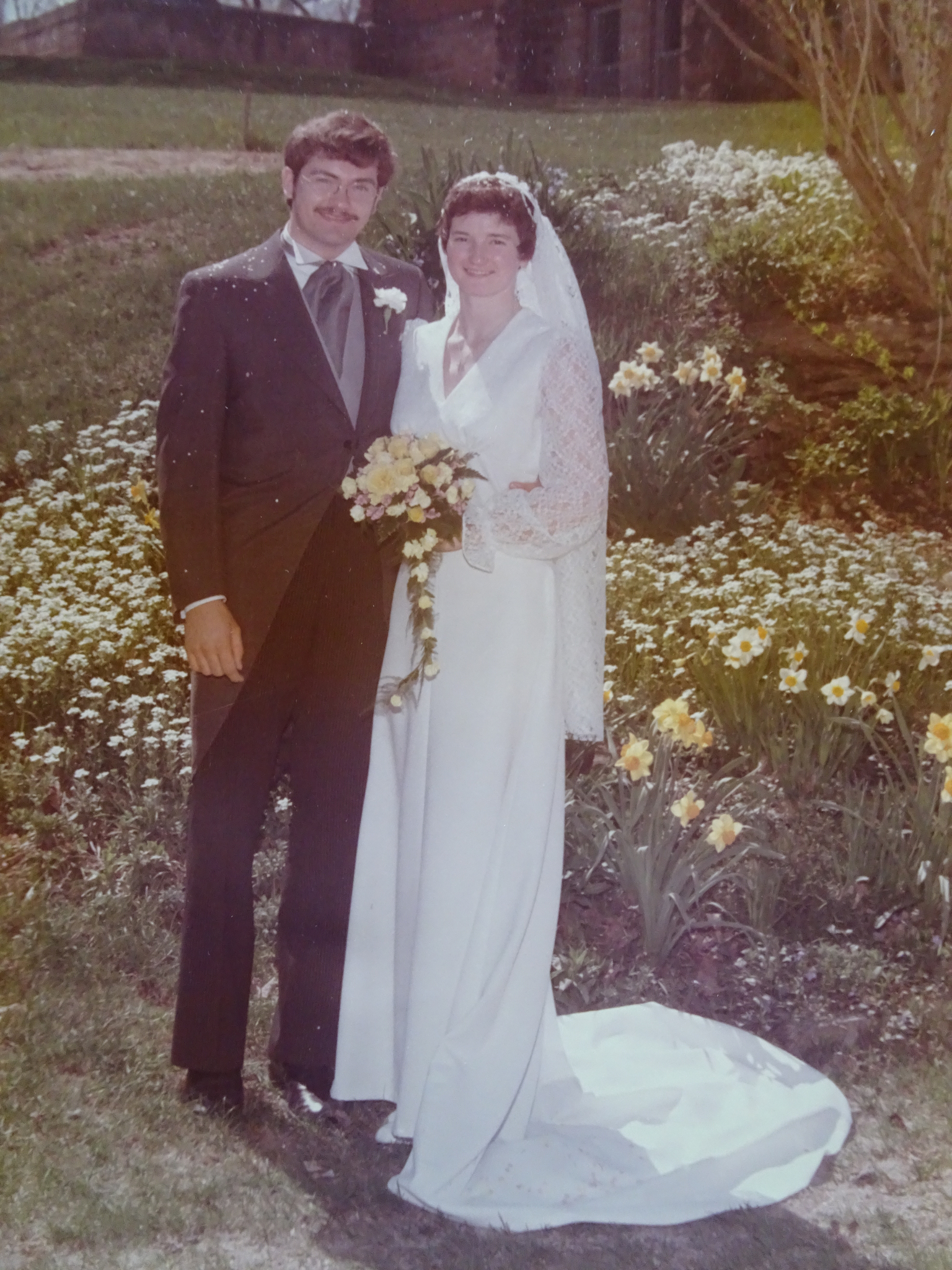 John and Donna Greenhorn smile in a wedding photo outside in front of flowers in a garden. They wear wedding attire and Donna holds a bouquet of flowers.