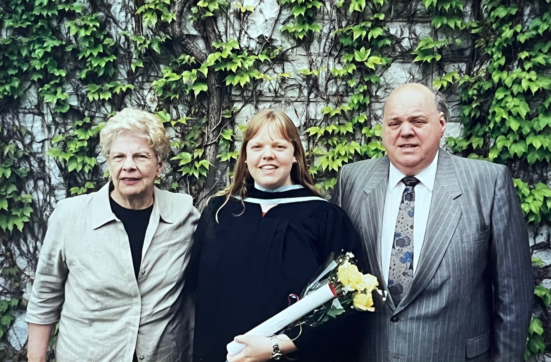 Julia Allen poses outside in Convocation robes with her parents in front of an ivy covered wall.