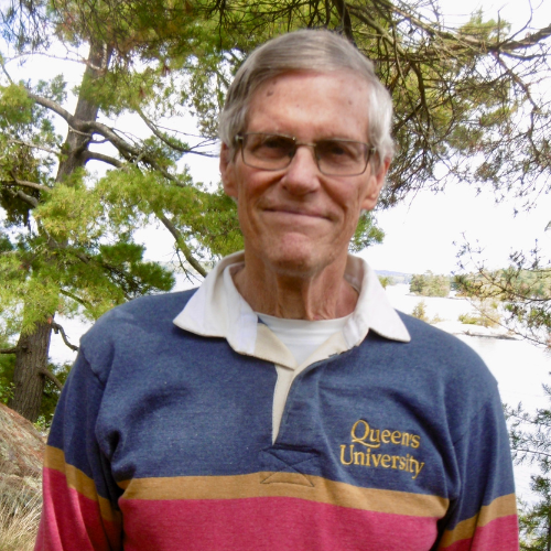 Len Minty wears a Queen's sweater and glasses outside in front of a body of water.