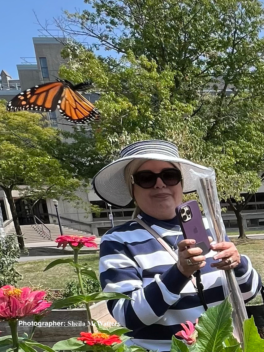 Maha smiles in the gardens wearing a hat and sunglasses. A butterfly flies in the foreground of the image.