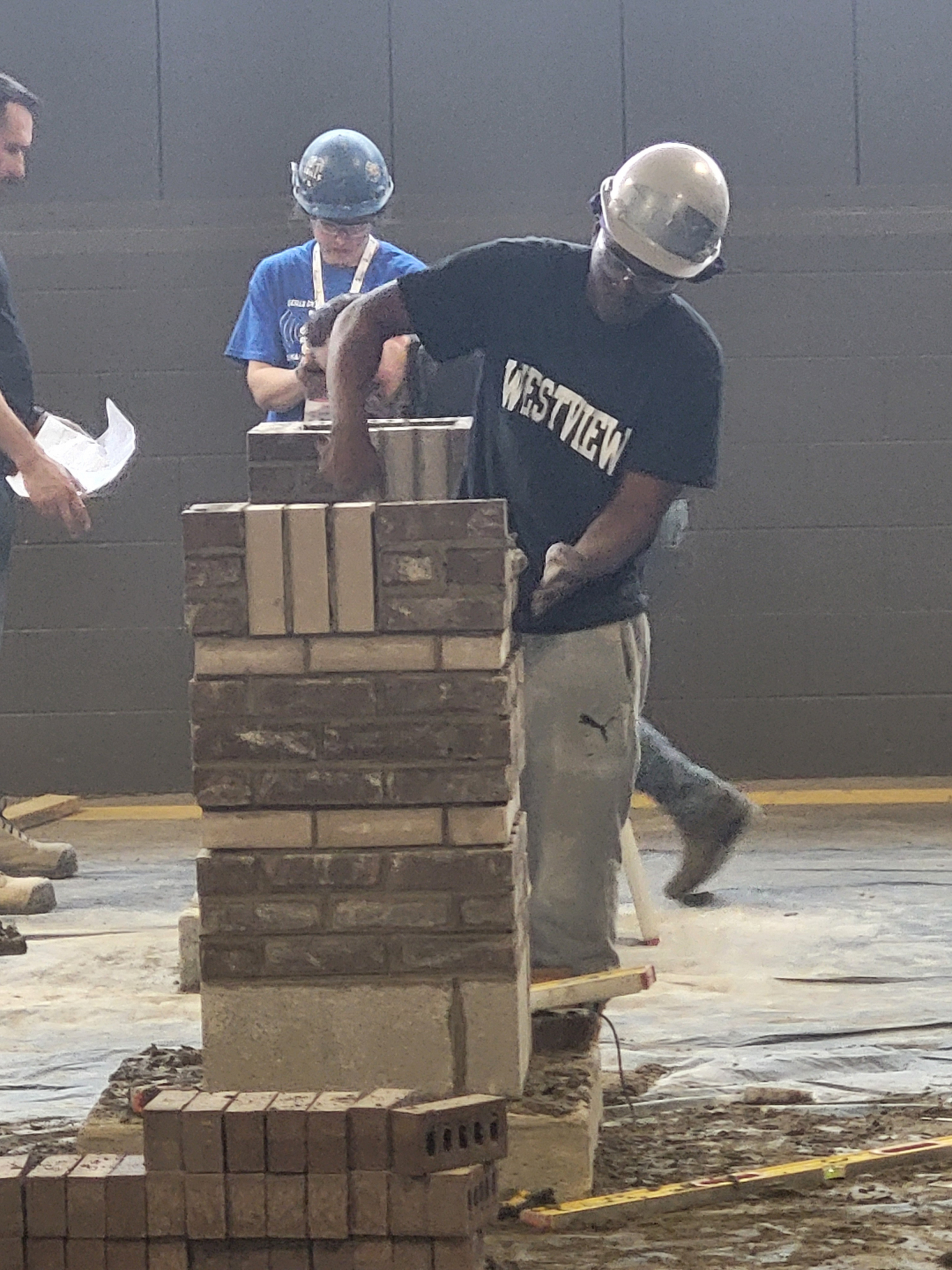 A student in a hard hat and safety goggles stands owrking on a brick masonry project. The student is in the middle of using a tool to apply more concrete.