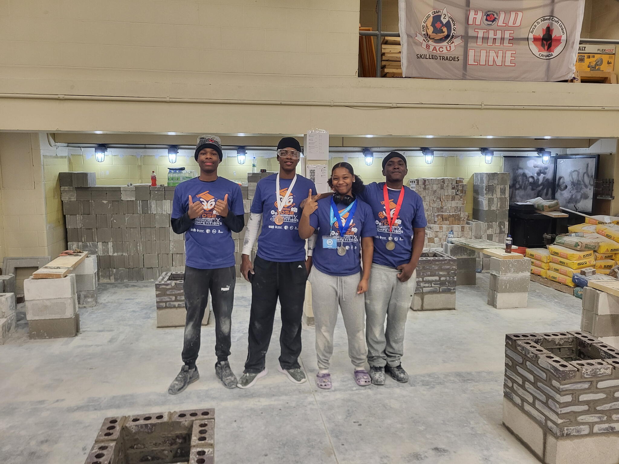 Four students pose in a tech classroom wearing matching shirts and three wear medals. They all smile at the camera.