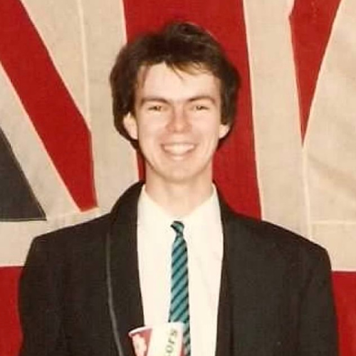 Michael Kehler during his time at the Faculty of Education with brown hair wearing a black suit jacket with a union jack flag behind him.