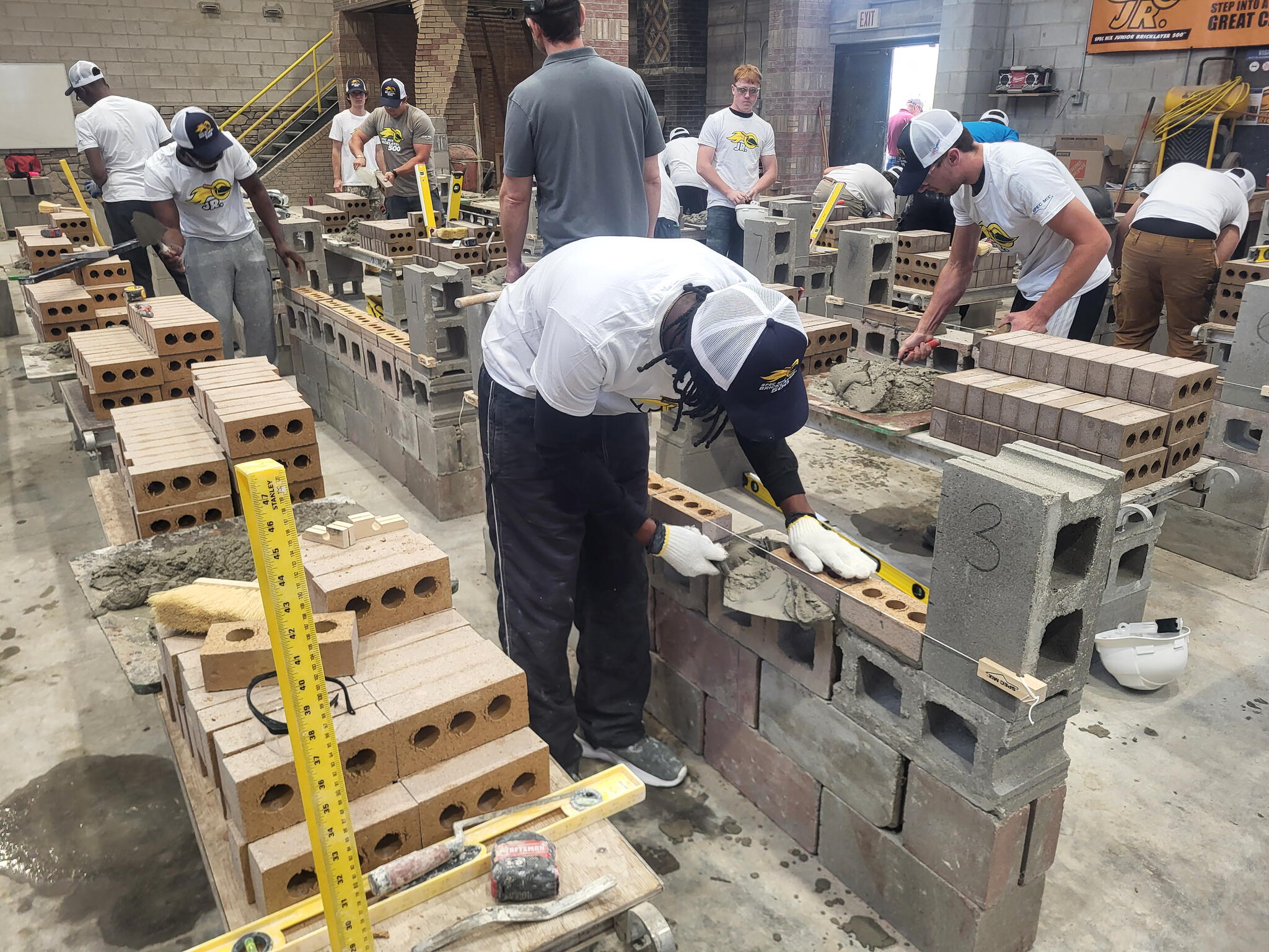 A student wears gloves and holds a trowel with material next to a brick wall. There are several other students working on projects in a tech classroom. They all wear tshirts and ball caps.
