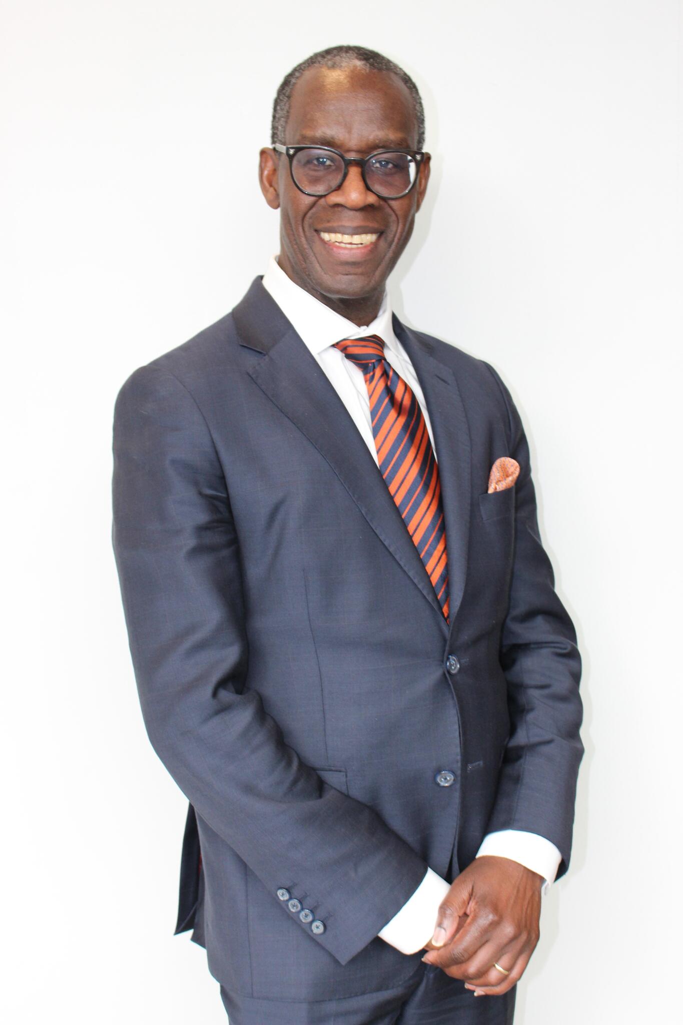 Opiyo Oloya smiles standing in front of a white wall holding his hands.