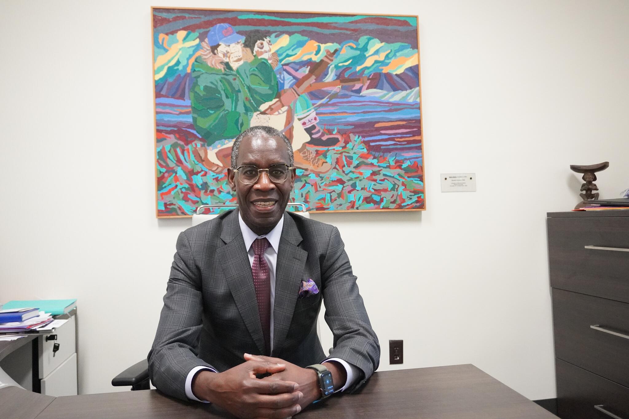 Opiyo Oloya smiles seated behind a desk with his hands clasped on the table. He wears glasses in front of a colourful painting.