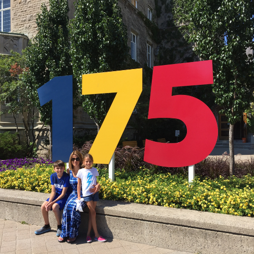 Rose Heron poses with her two children in front of a Queen's anniversary sign that reads "175."