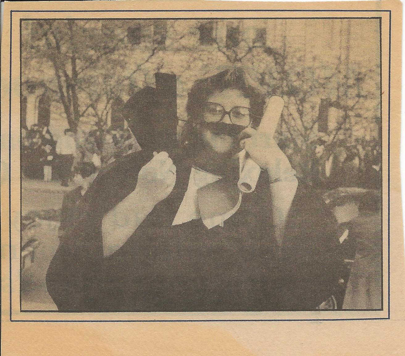 Sheri Henderson smiles in a photo in a newspaper. Sheri is at graduation holding a degree and cap and wearing a gown. She holds up part of the cap to make a mustache.