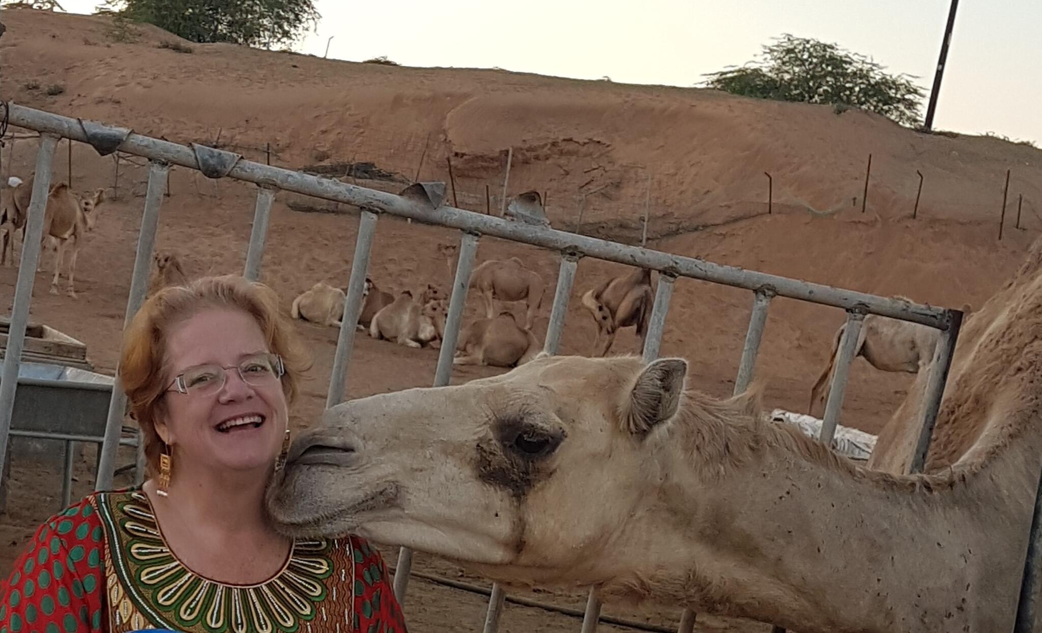 Sheri Henderson looks towards the camera while a camel sniffs her neck.