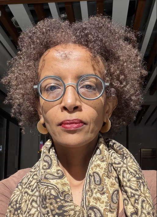 Tigist Amdemichael smiles wearing glasses, red lipstick, and scarf and poses in front of the light from a window.