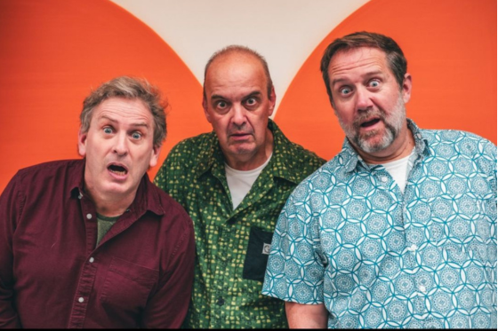 The band The Arrogant Worms poses in front of a graphic orange and white wall. The band members make surprised expressions and look intently at the camera.