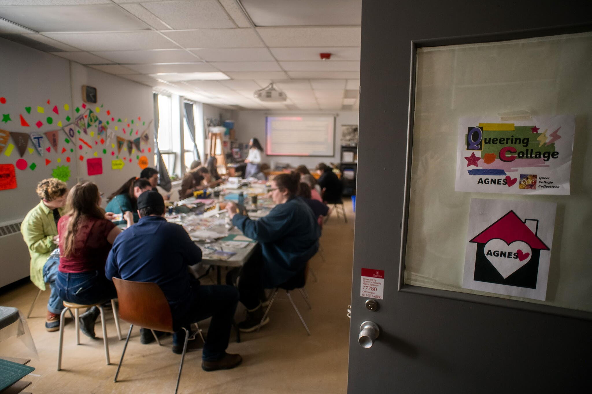 A classroom like space filed with people creating art with posters on the classroom door saying AGNES and Queering Collage
