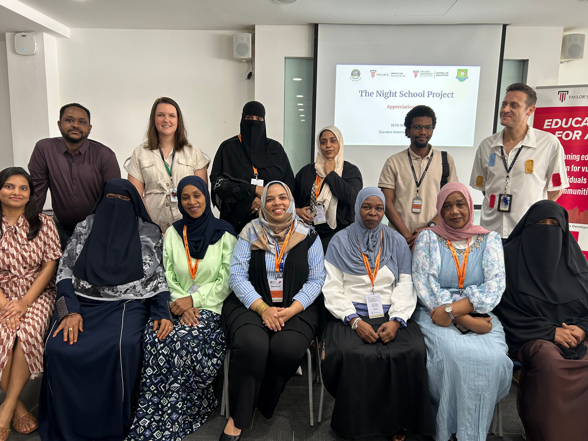 A group of educators in burkhas and headscarves smile with instructors at a certificate ceremony. A screen in the back reads "The Night School Project."