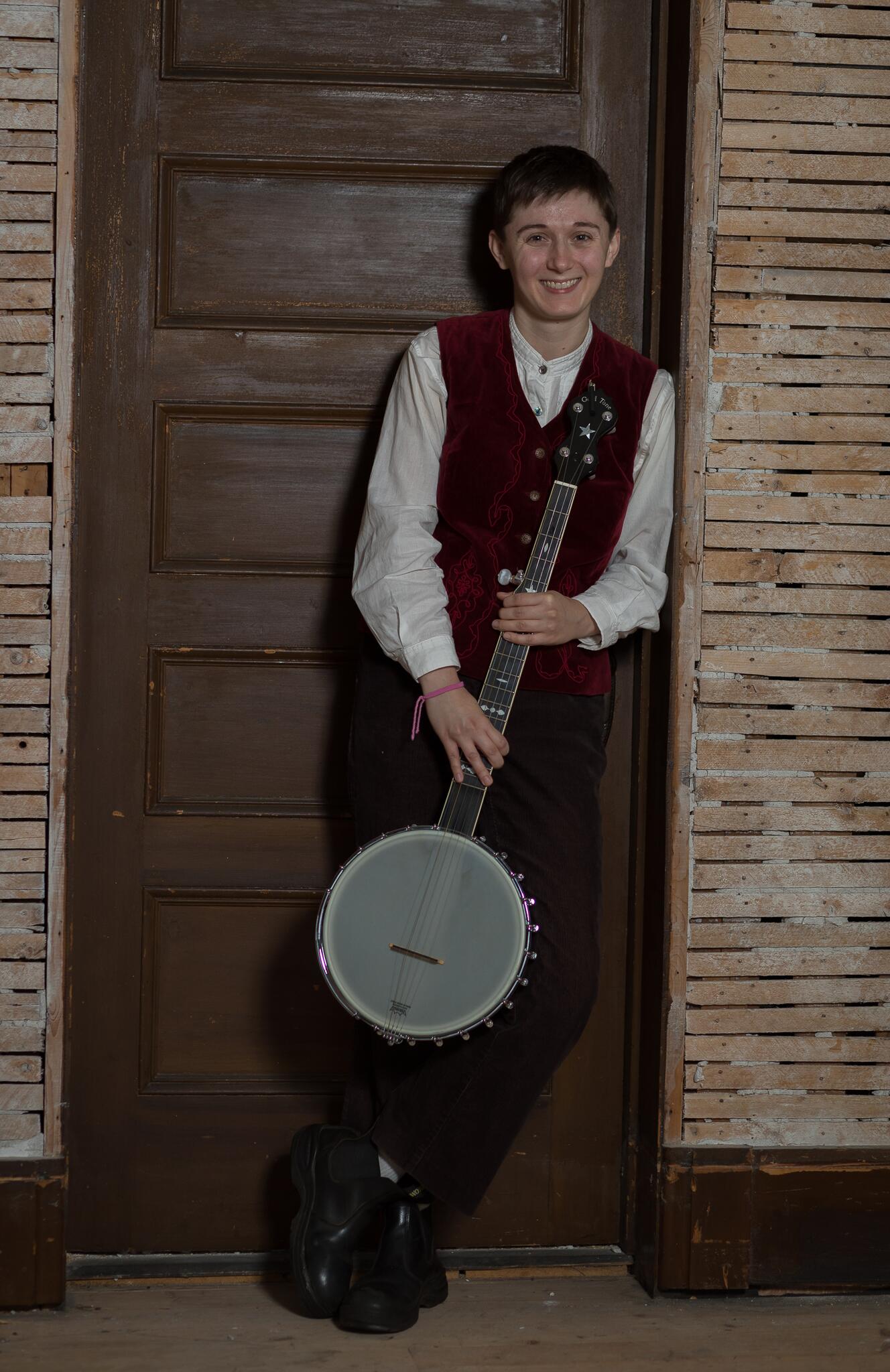 Anthea Feaver smiles leaning against a door frame and resting a banjo against her that she holds pointing towards the ground in both hands. She wears a red velvet vest and white long sleeve shirt.