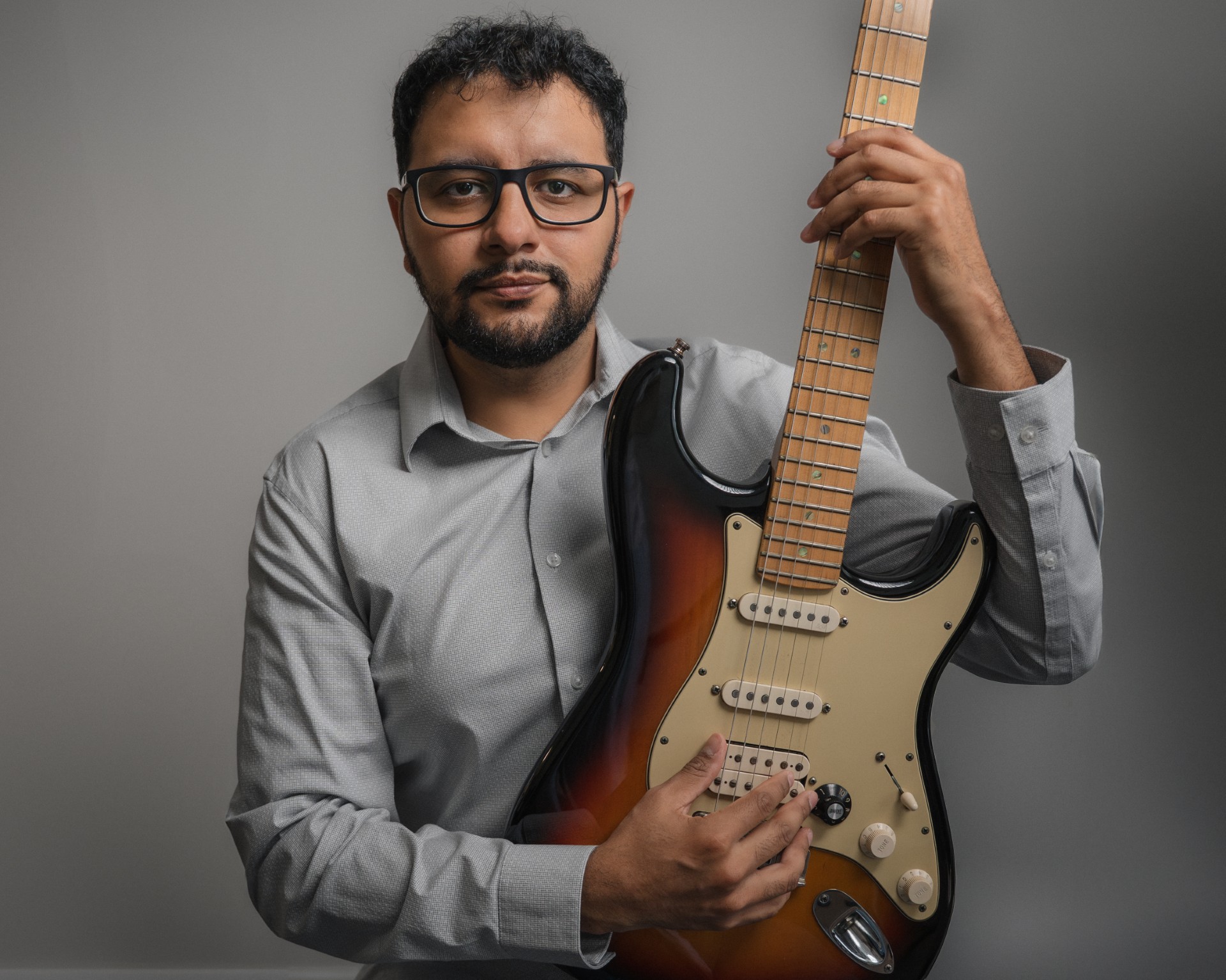 Juan Velasco looks at the camera and holds a guitar. He sits in front of a grey background wearing a grey collared shirt.